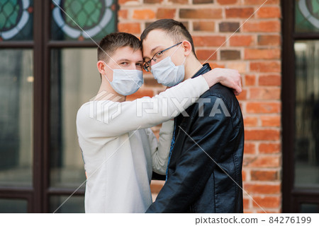 Young gay couple wearing medical mask, hugging and kissing at the city. Young gay couple wearing medical mask, hugging and kissing at the city. 84276199