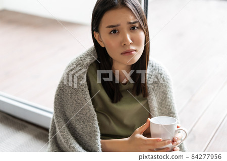Sad and gloomy asian woman sitting at home near window, holding cup of coffee and frowning disappointed, feeling unhappy or miserable Sad and gloomy asian woman sitting at home near window, holding cup of coffee and frowning disappointed, feeling unhappy or miserable 84277956