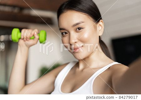 Portrait of smiling asian fitness instructor taking selfie with dumbbell. Athletic woman making photo on mobile phone with sport gear, workout at home 84279017