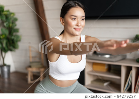Young asian fitness woman with healthy fit body, doing squats, morning workout, wearing activewear, standing at home in living room and having training session Young asian fitness woman with healthy fit body, doing squats, morning workout, wearing activewear, standing at home in living room and having training session 84279257
