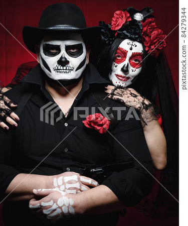 beautiful woman with a sugar skull makeup with a wreath of flowers on her head and a skeleton man in a black hat. Couple on dark red background beautiful woman with a sugar skull makeup with a wreath of flowers on her head and a skeleton man in a black hat. Couple on dark red background 84279344