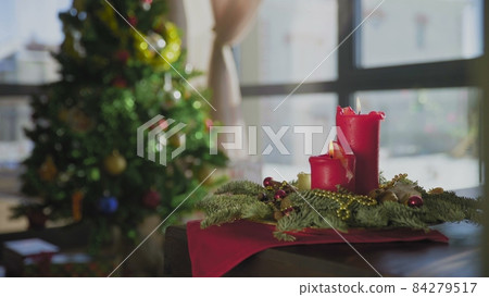 An evergreen wreath with two red candles and one golden candle. In the background, a decorated Christmas tree. Christmas traditions and customs of Christians on the eve of the holiday. An evergreen wreath with two red candles and one golden candle. In the background, a decorated Christmas tree. Christmas traditions and customs of Christians on the eve of the holiday. 84279517