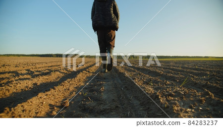 Low angle: man walking in rubber boots in a farmer's field, the blue sky above the horizon. Man walking through an agricultural field. Farmer walks through a plowed field in early spring. 84283237