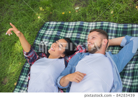 Couple in love resting in park on plaid or blanket. Girl is showing something boyfriend. Handsome guy and pretty girl watching at the sky Couple in love resting in park on plaid or blanket. Girl is showing something boyfriend. Handsome guy and pretty girl watching at the sky 84283439