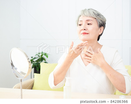 Happy 50s middle aged asian woman touching face skin looking in mirror reflection. Smiling mature old lady pampering, healthy moisturized skin care, aging beauty, skincare treatment cosmetics concept. 84286366