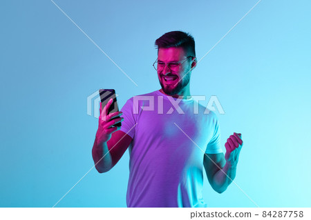 Cropped portrait of happy excited man joyfully looking at phone screen isolated over blue background in neon lights 84287758