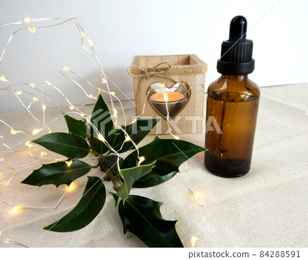Cosmetic oil transparent glass brown bottle with holly plant, candle and decorative illumination garland, simple minimalist Christmas holiday still life arrangement, eco-friendly concept 84288591