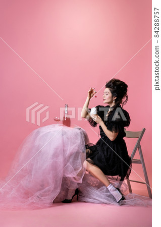 Young pretty woman with retro style hairdo in black evening little dress sitting at holiday table isolated on pink background. 84288757