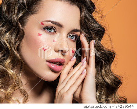 Portrait of a beautiful girl with painted red hearts on her face. Fashion photography. Sexy blonde with curly hair. An expressive and sensual model with bright makeup poses in the studio. Portrait of a beautiful girl with painted red hearts on her face. Fashion photography. Sexy blonde with curly hair. An expressive and sensual model with bright makeup poses in the studio. 84289544