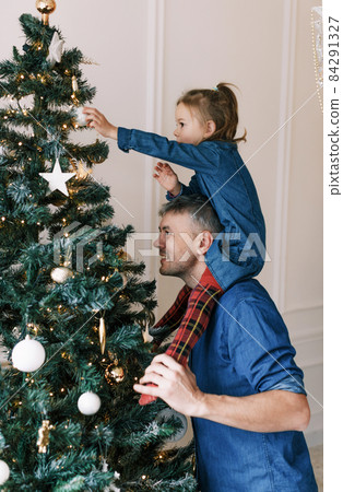 Happy dad and daughter decorate the Christmas tree. A little girl is sitting on Daddy's shoulders, holding a toy on the Christmas tree. happy christmas 84291327