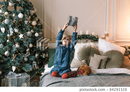 a little beautiful girl is sitting on the bed next to the Christmas tree holding a gift high up 84291328
