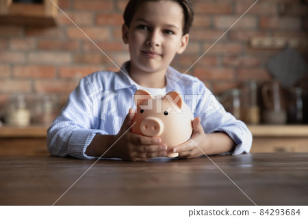 Preteen boy stand at kitchen table hold piggybank save money Preteen boy stand at kitchen table hold piggybank save money 84293684