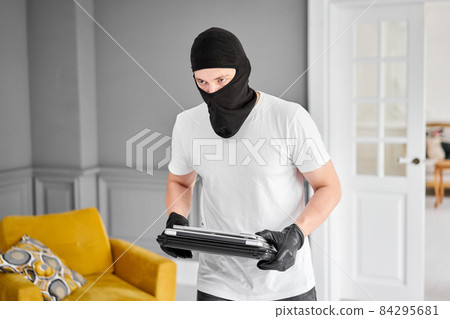 Thief with black balaclava stealing modern expensive laptop from apartment. Masked face. Man burglar stealing tv set from house 84295681