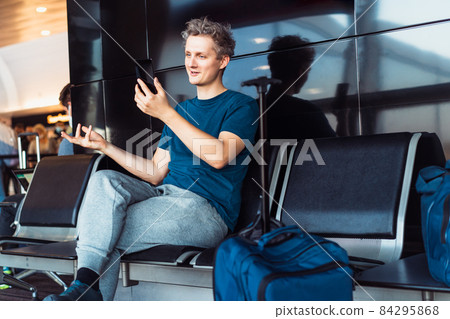 Young male traveler waiting for his flight in the airport and having video meeting. call, chat by his smartphone. Digital nomad, freelance lifestyle. Selective focus. 84295868