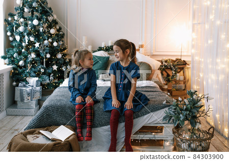 two little girls are sitting on the edge of a large bed looking at each other against the background of a Christmas tree. happy christmas 84303900