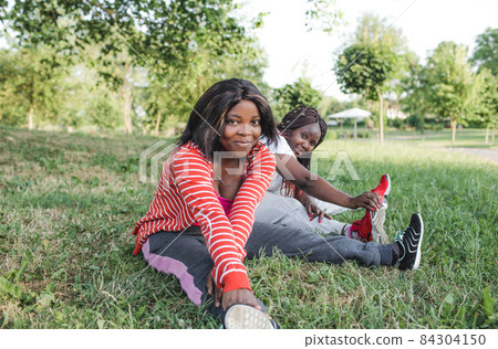 Two African-American girls are engaged in sports, doing exercises sitting in the park on the grass. 84304150