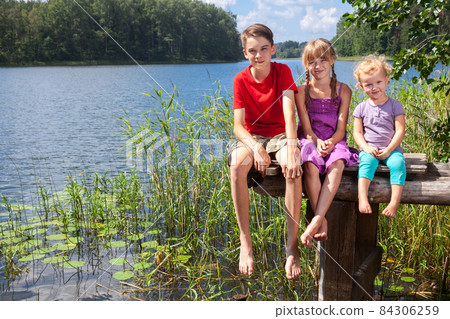 Mixed age children sitting on a pier by a summer lake Mixed age children sitting on a pier by a summer lake 84306259