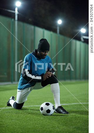 Young African footballer standing on knee on football field Young African footballer standing on knee on football field 84307064