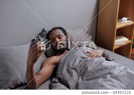 Young man looking at alarm clock in his hand after awakening Young man looking at alarm clock in his hand after awakening 84307228