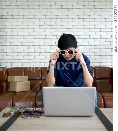 Young man wearing sunglasses facing to computer screen..Asian man sell various styles of eyeglasses online via application on laptop computer. Young man wearing sunglasses facing to computer screen..Asian man sell various styles of eyeglasses online via application on laptop computer. 84307873