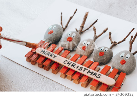 Merry Christmas deer on greeting card postcard made of pebbles, sea stones, clothespins and branches on white background. Diy Natural Eco style diy. Gift idea. Step by step. Top view. Merry Christmas deer on greeting card postcard made of pebbles, sea stones, clothespins and branches on white background. Diy Natural Eco style diy. Gift idea. Step by step. Top view. 84309352