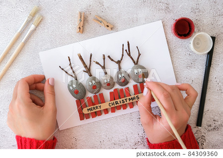 Diy Merry Christmas deer on greeting card postcard made of pebbles, sea stones, clothespins and branches on white background. Natural Eco style diy. Gift idea. Step by step. Top view. Diy Merry Christmas deer on greeting card postcard made of pebbles, sea stones, clothespins and branches on white background. Natural Eco style diy. Gift idea. Step by step. Top view. 84309360