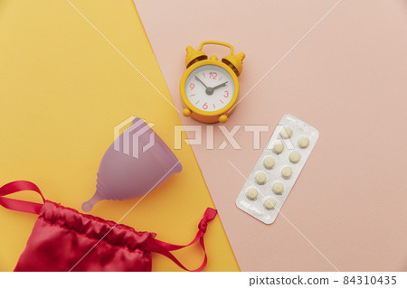 Top view of sanitary cup with pain relief pills and an alarm clock Top view of sanitary cup with pain relief pills and an alarm clock 84310435