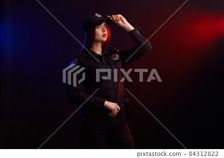 Serious female police officer is posing for the camera against a black background with red and blue backlighting. Serious female police officer is posing for the camera against a black background with red and blue backlighting. 84312822