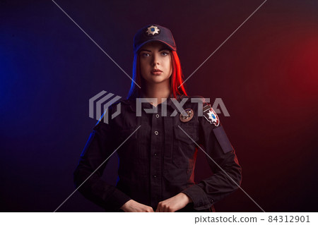 Serious female police officer is posing for the camera against a black background with red and blue backlighting. Serious female police officer is posing for the camera against a black background with red and blue backlighting. 84312901