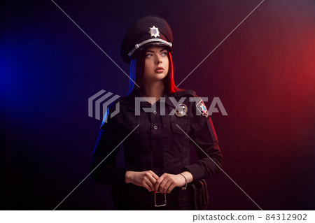 Serious female police officer is posing for the camera against a black background with red and blue backlighting. Serious female police officer is posing for the camera against a black background with red and blue backlighting. 84312902