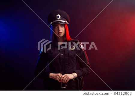 Serious female police officer is posing for the camera against a black background with red and blue backlighting. Serious female police officer is posing for the camera against a black background with red and blue backlighting. 84312903