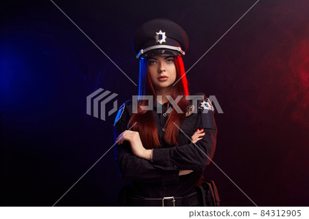 Serious female police officer is posing for the camera against a black background with red and blue backlighting. Serious female police officer is posing for the camera against a black background with red and blue backlighting. 84312905