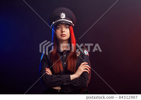 Serious female police officer is posing for the camera against a black background with red and blue backlighting. Serious female police officer is posing for the camera against a black background with red and blue backlighting. 84312907
