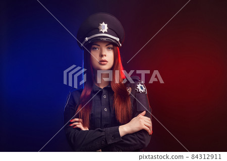 Serious female police officer is posing for the camera against a black background with red and blue backlighting. Serious female police officer is posing for the camera against a black background with red and blue backlighting. 84312911