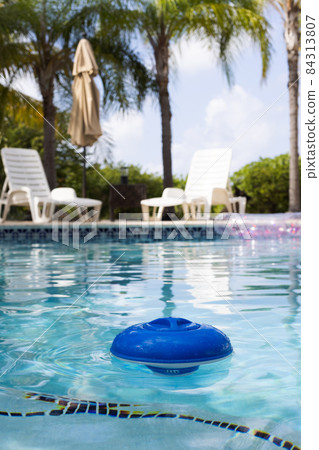 Blue plastic chlorine dispenser floating on the surface of a swimming pool surrounded by palm trees. Blue plastic chlorine dispenser floating on the surface of a swimming pool surrounded by palm trees. 84313807