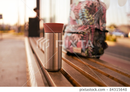 Close Up shot of a trip hiking bag with metal thermos mug on bench at bus stop. Train station. Close Up shot of a trip hiking bag with metal thermos mug on bench at bus stop. Train station. 84315648