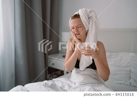 Sad unhappy europian girl with toothache holds a glass of clean water in a white bed of a bed at home in the morning 84315818