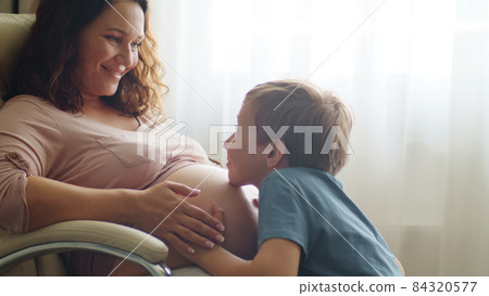 Child hugs her mother's pregnant belly. Happy son strokes the big belly his pregnant mom near window at home. Happy family life in an expectation a newborn. Pregnancy and maternity. 84320577