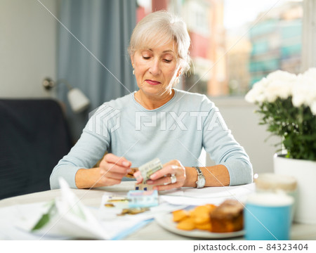Elderly woman sitting at table with papers and counting money Elderly woman sitting at table with papers and counting money 84323404