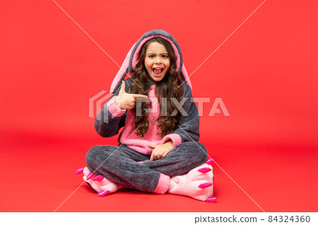 amazed kid in comfortable pajama sit pointing finger on herself on red background, morning 84324360