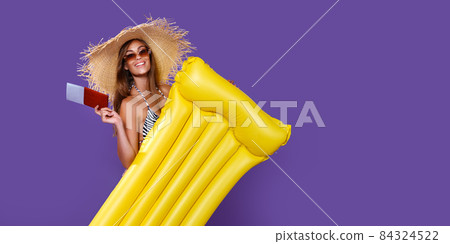 Smiling girl in bikini going to travel stands with yellow inflatable mattress holds ticket, passport 84324522