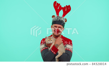 Lovely adult man in fashionable Christmas sweater and deer antlers hat holding his beloved cat pet Lovely adult man in fashionable Christmas sweater and deer antlers hat holding his beloved cat pet 84328014