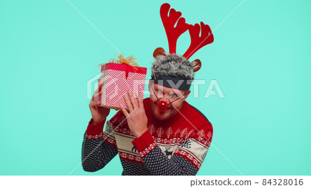 Funny man wears New Year sweater deer antlers received present, interested in what inside gift box Funny man wears New Year sweater deer antlers received present, interested in what inside gift box 84328016