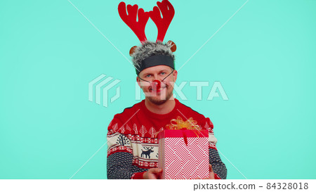 Funny man wears red New Year sweater and deer antlers presenting Christmas gift box, shopping sale Funny man wears red New Year sweater and deer antlers presenting Christmas gift box, shopping sale 84328018