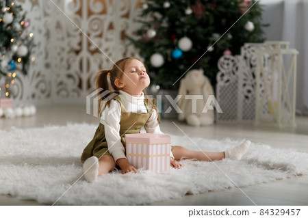 Offended little girl on a white carpet with new year gift against the background of Christmas trees Offended little girl on a white carpet with new year gift against the background of Christmas trees 84329457