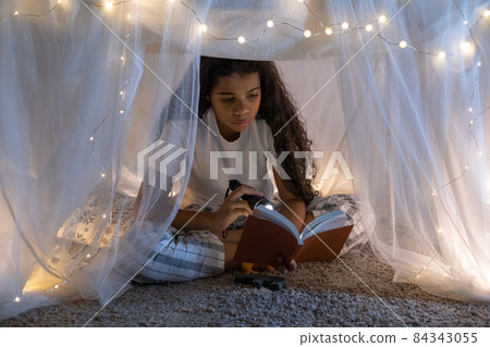 Concentrated wavy-haired African American girl in pijamas sitting with crossed legs inside handmade tent and reading book with flashlight Concentrated wavy-haired African American girl in pijamas sitting with crossed legs inside handmade tent and reading book with flashlight 84343055