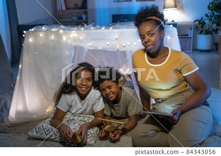 Portrait of careful African American mother with tablet sitting at handmade tent and reading fairy tale to kids 84343056