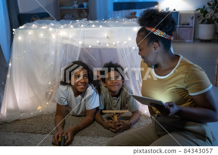 Smiling blackwoman using tablet while reading online book to children inside of play tent before bed Smiling blackwoman using tablet while reading online book to children inside of play tent before bed 84343057