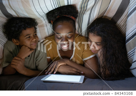 Smiling young African American mother lying on front and reading book to children under duvet 84343108