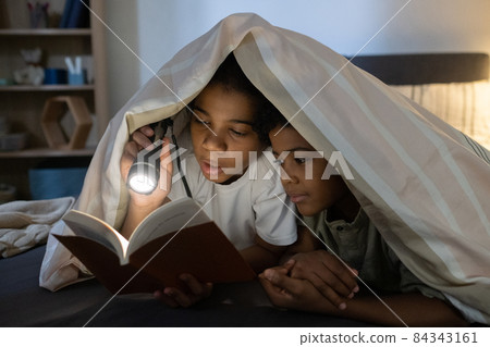 African American girl reading book to younger brother under light of flashlight while they lying under duvet in dark room African American girl reading book to younger brother under light of flashlight while they lying under duvet in dark room 84343161
